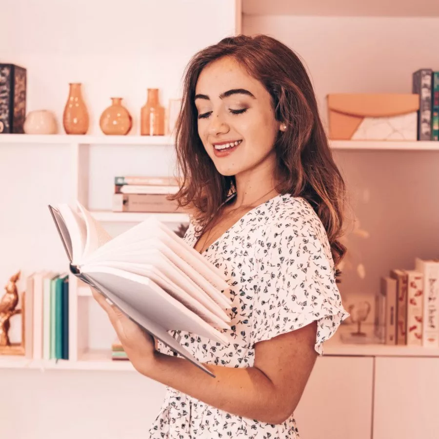 Author Abbie Emmons smiles while reading a book. The background is a monochromatic coral pink room, delicate and pastel in feel.